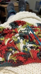 Image shows a blue crochet hook in front of mult-colored crochet in progress, about 9 inches deep. It is on top of a purchased white knitted throw. This is the “right” side.