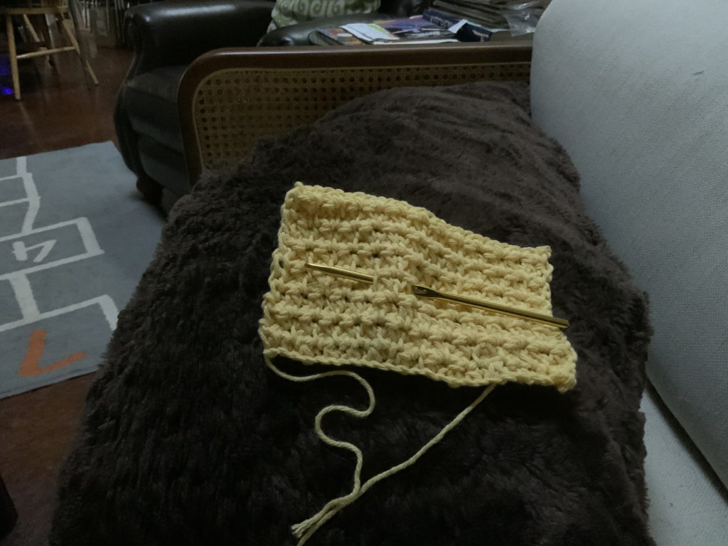 test swatch of yellow cotton yarn with crochet hook on top of dark brown pillow.