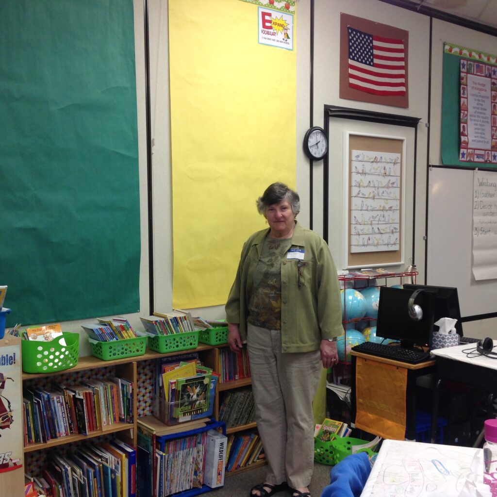 my mother standing in my original third grade classroom, in front of a wall of posters, books, and anchor charts.